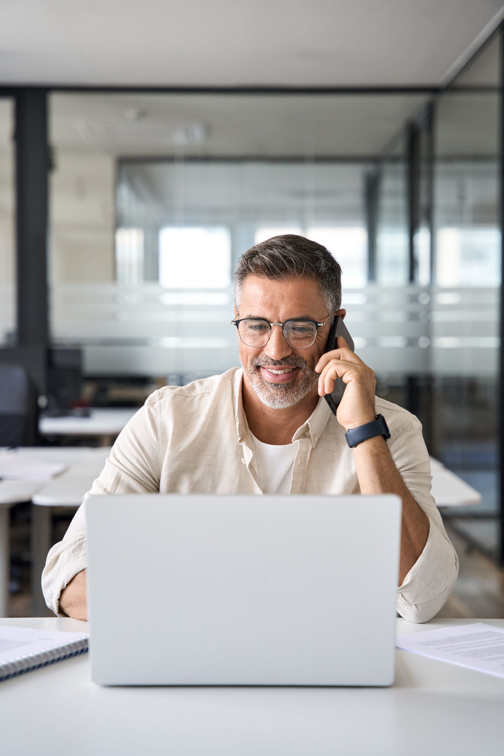 Middle aged Latin or Indian businessman having call on smartphone with business partner or client. Vertical portrait of smiling mature Hispanic man sitting at table talking by mobile cellphone indoors